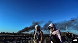 Transeúntes caminan por un puente en Leópolis, en el oeste de Ucrania, mientras una columna de humo se eleva en la distancia tras una explosión cerca del aeropuerto de la ciudad el 18 de marzo de 2022.