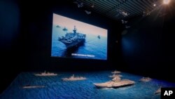 A TV screen showing the U.S. Navy fleet sail in formation near the models of Liaoning aircraft carrier with navy frigates and submarines on display at the military museum in Beijing, Aug. 1, 2019.